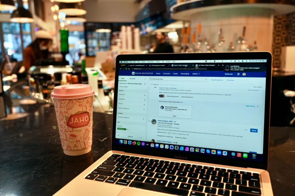 An open MacBook Air sits on a dark counter inside a cafe, displaying a LinkedIn Sales Navigator interface on its screen. Next to the laptop stands a disposable Jaho Artisan Coffee cup with a pink lid, set against a softly blurred background of the cafe's interior.