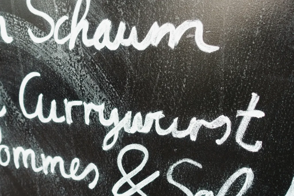 A close-up shot shows German menu items handwritten in white chalk on a black chalkboard. The clearly visible text prominently features the word "Currywurst" above "Pommes & Sal...".