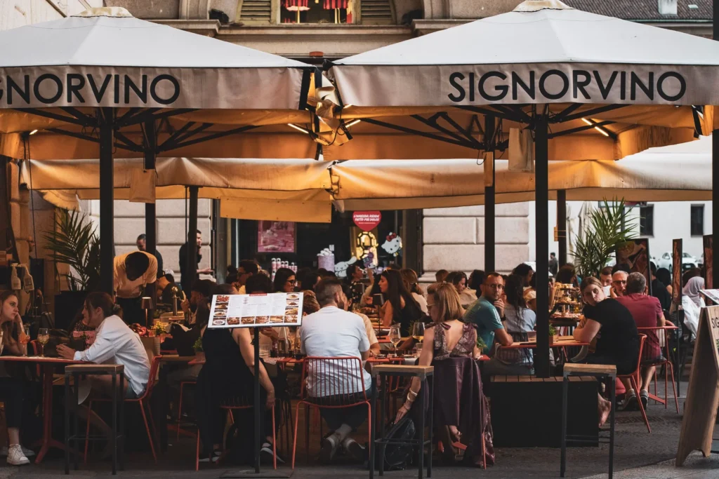 An outdoor seating area of a Signorvino restaurant is filled with patrons dining under large beige umbrellas. People sit at small tables enjoying food and drinks in a lively urban setting.