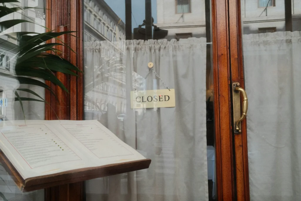 A closed sign hangs in the window of an elegant restaurant behind a sheer white curtain. In the foreground, an open menu rests on a wooden stand outside the glass door.