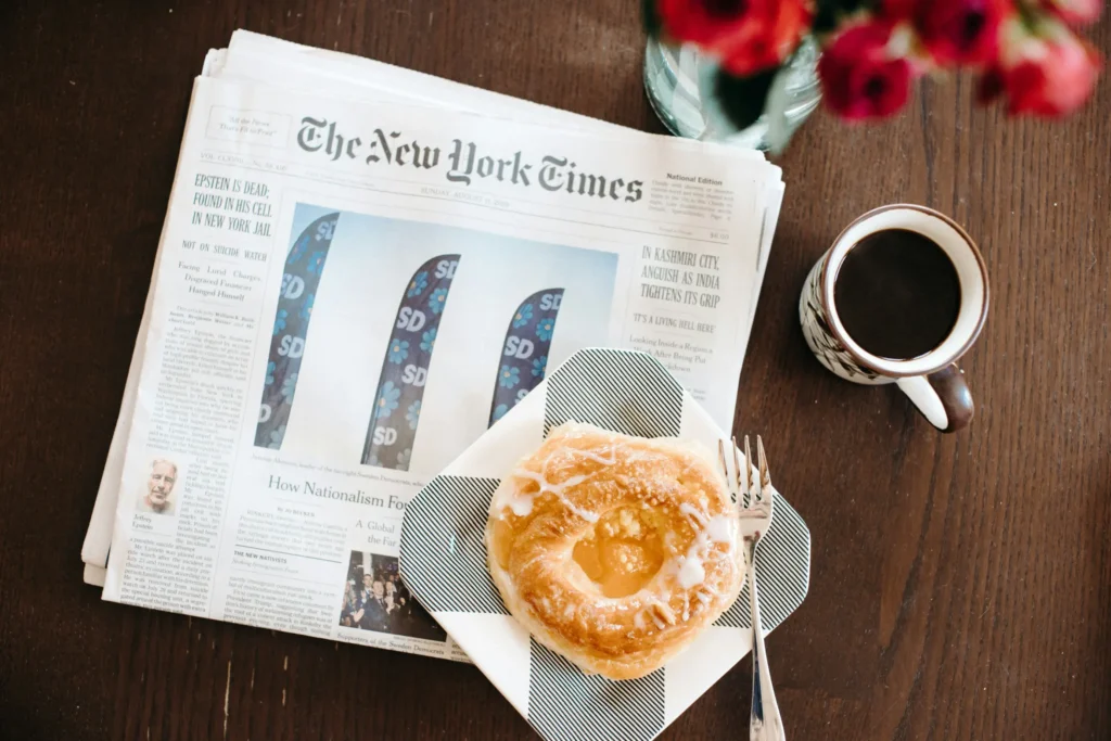 A top-down view shows a copy of The New York Times newspaper laid out on a dark wooden table next to a cup of black coffee. Resting on the front page is a small, patterned plate holding a glazed pastry and a silver fork, with red flowers subtly visible at the top.