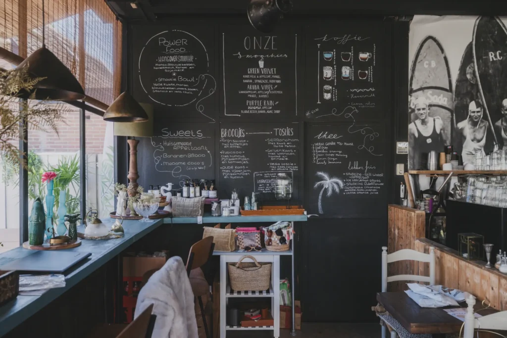 A cozy café interior features a large black chalkboard wall covered in handwritten menus for coffee, smoothies, and light bites. Hanging pendant lights illuminate a long wooden counter decorated with glassware, set alongside a vintage black-and-white surfer mural on the far right.