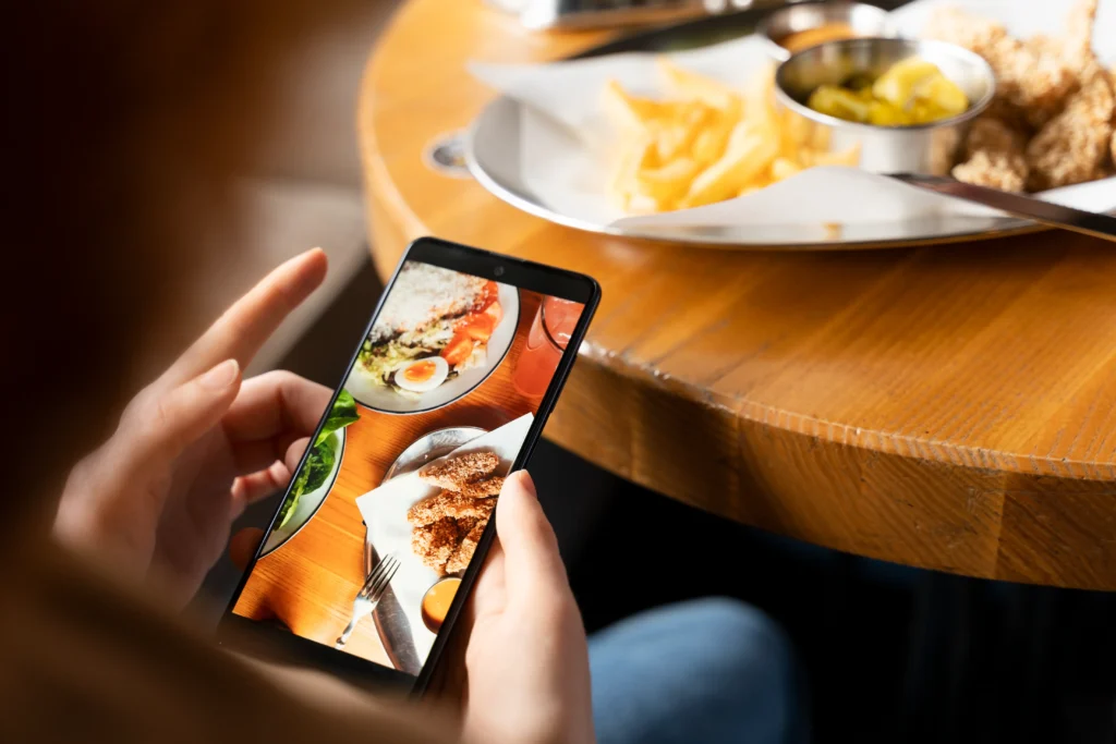A person holds a smartphone, displaying a photo of a restaurant meal with salad and fried chicken tenders. In the background, a plate of french fries, dipping sauces, and chicken sits on a wooden table.