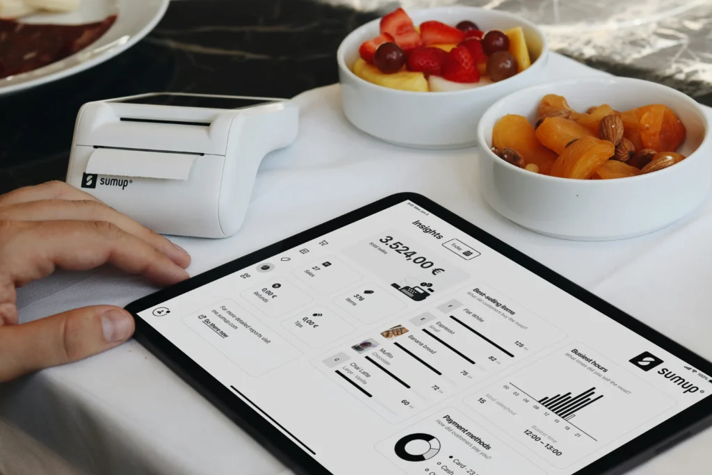 A tablet displaying a SumUp point-of-sale analytics dashboard rests on a white tablecloth next to a small receipt printer. In the background, ceramic bowls filled with fresh fruit, dried apricots, and nuts sit on the table.