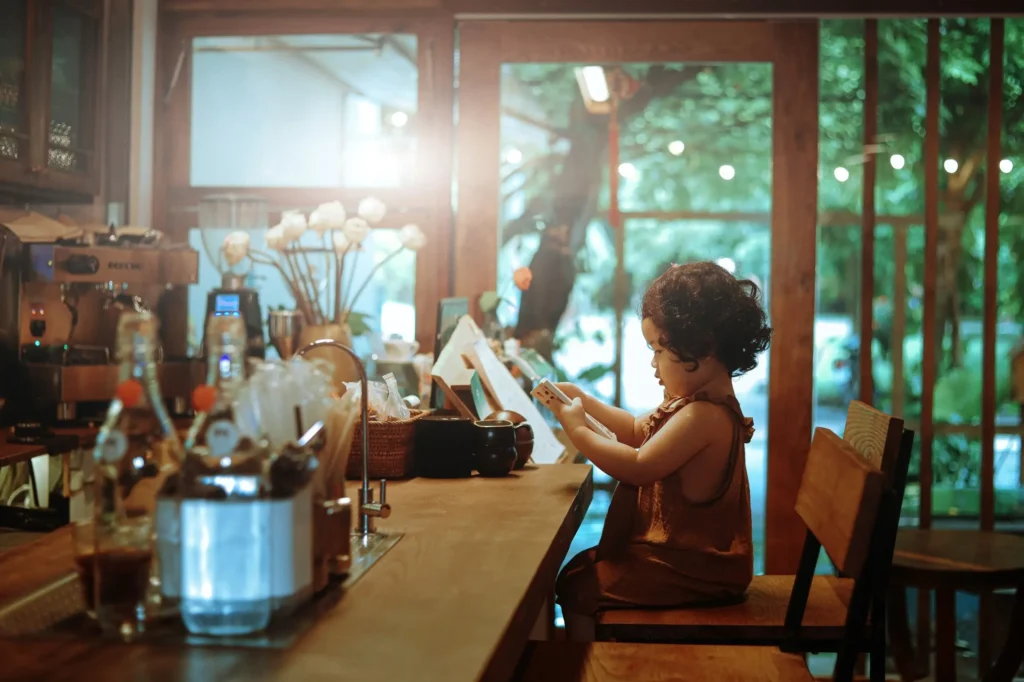 A young child with curly dark hair sits at a wooden counter inside a warmly lit cafe, holding and examining a white smartphone. The background features coffee-making equipment, decorative flowers, and large glass windows looking out onto lush greenery.