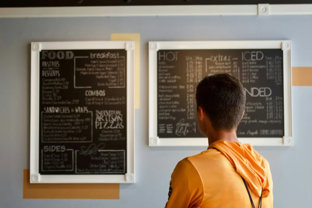 A young man in an orange hoodie stands facing two framed chalkboard menu boards in a café. He appears to be reading through the listed food and drink options while deciding what to order.