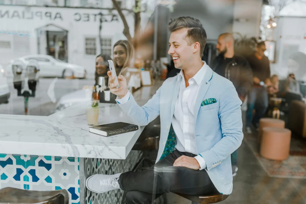 A stylishly dressed young man wearing a light blue blazer smiles as he looks at his smartphone while seated at a bar counter. Next to him on the tiled counter sit a iced drink and a notebook, with people and street views visible in the background through the glass window.