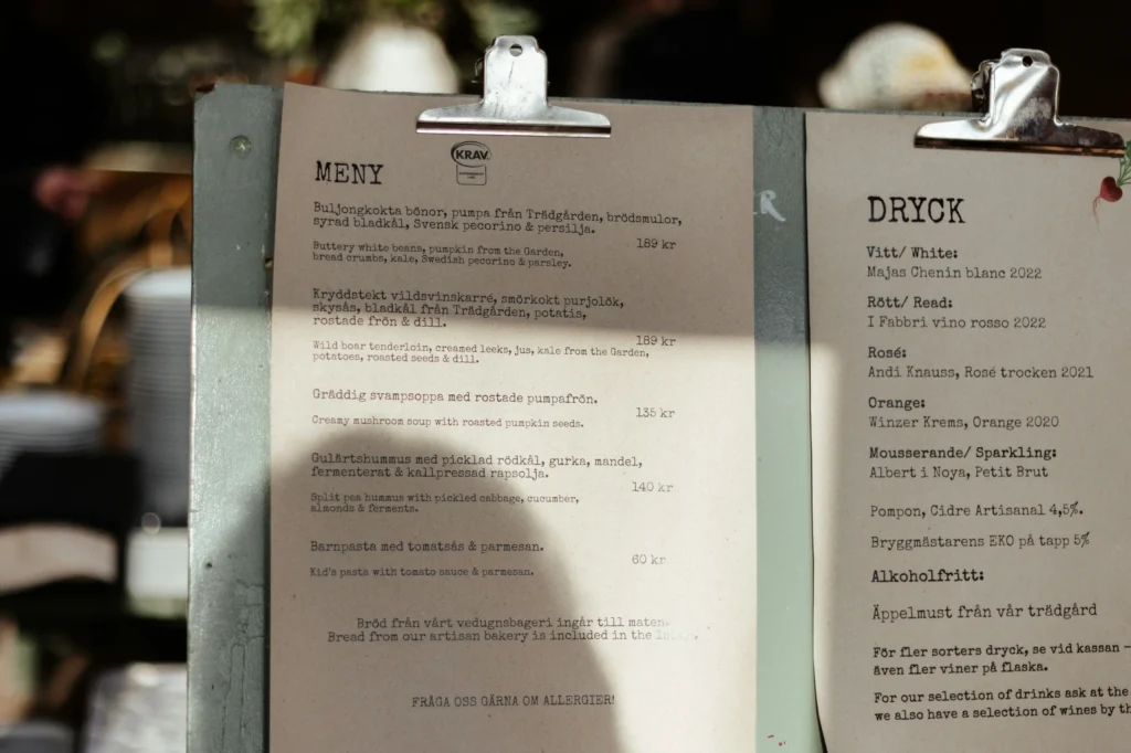 A close-up shot of two beige paper menu pages attached to metal clipboards, featuring Swedish and English text listing food items on the left and drinks on the right. Warm sunlight casts shadows across the printed food menu ("MENY") and beverage menu ("DRYCK"), highlighting items like wild boar tenderloin and various wines.