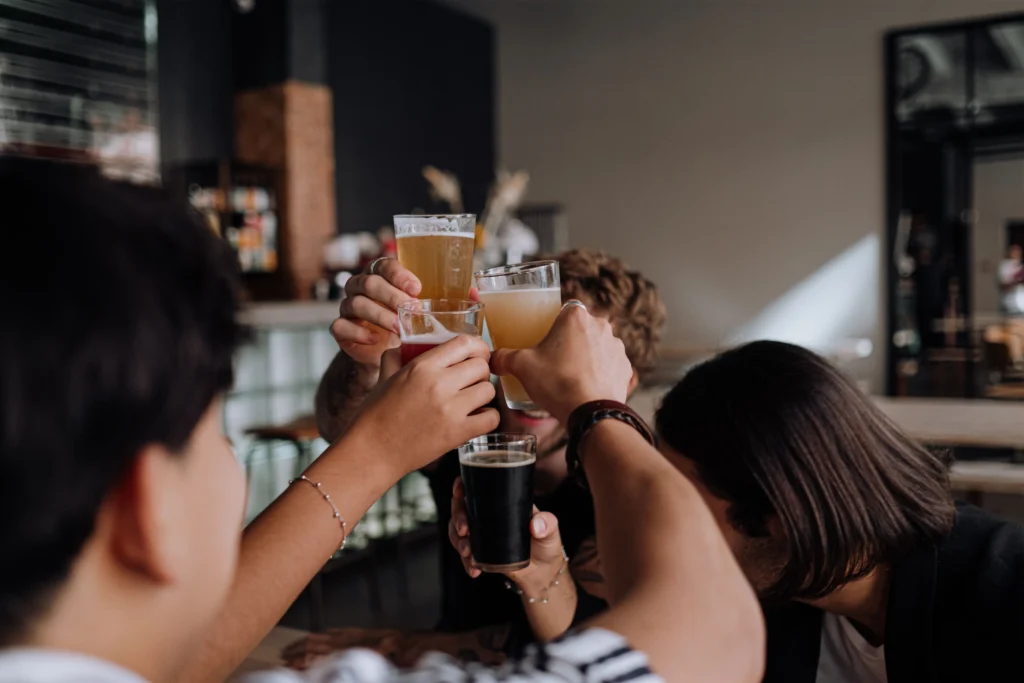 A group of friends raise their glasses in a toast together at a brightly lit bar table. The raised glasses hold a variety of drinks, including light beers, cider, and a dark stout.