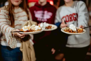 Three young people stand side-by-side, each holding out a paper plate piled with warm desserts topped with whipped cream and drizzle. The focus is on the sweet treats in the foreground, with the people blurred slightly in the background.