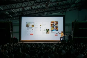 A speaker stands on a stage next to a large presentation screen displaying mobile application user interfaces. A seated audience watches the presentation in a spacious, dimly lit venue.