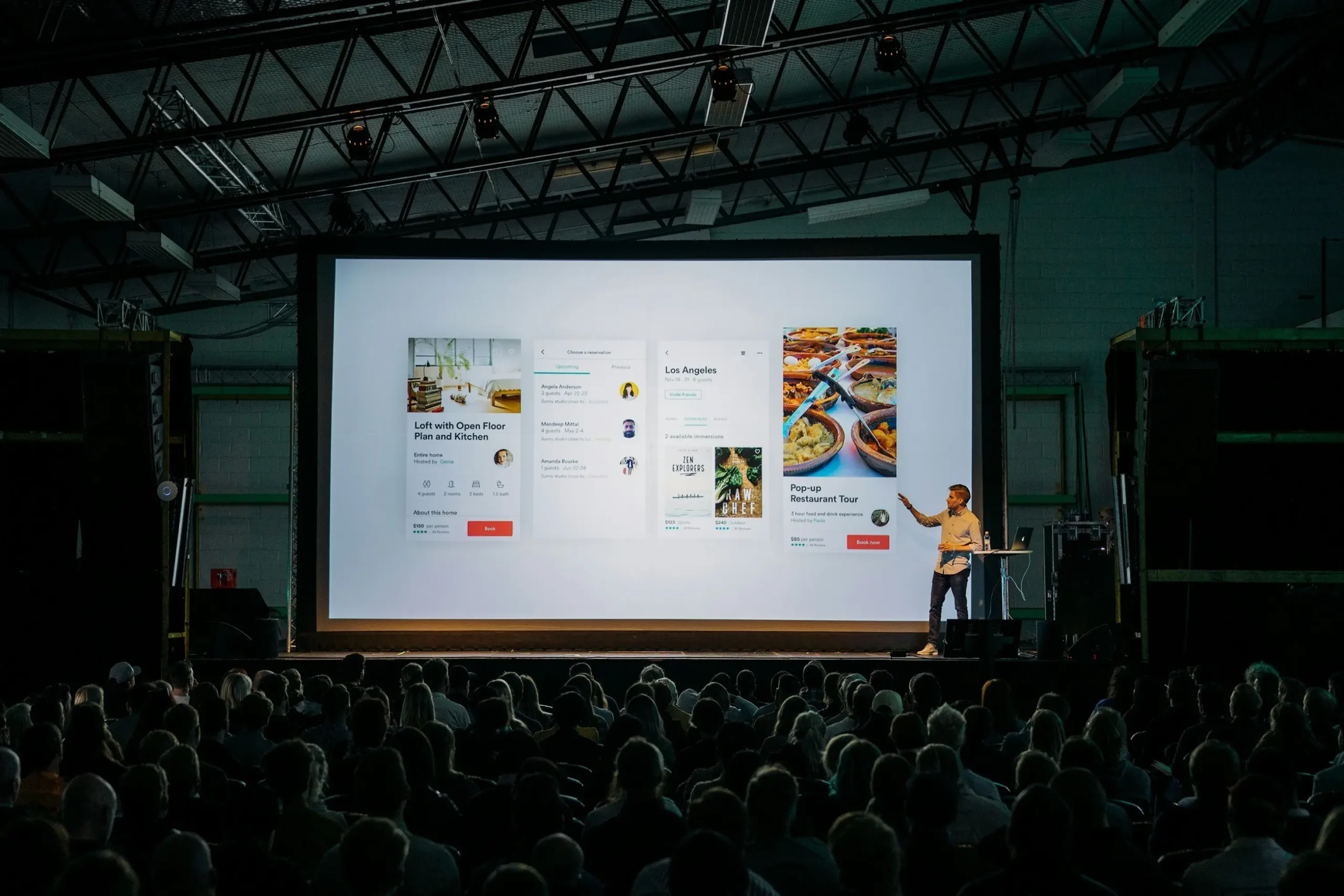 A speaker stands on a stage next to a large presentation screen displaying mobile application user interfaces. A seated audience watches the presentation in a spacious, dimly lit venue.