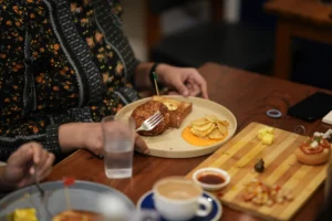 A person wearing a black patterned shirt holds a fork to eat a meal featuring a fried cutlet, toast, and potato chips served on a round plate. The surrounding wooden table is set with various breakfast dishes, including coffee, a wooden serving board with snacks, and glasses of water.