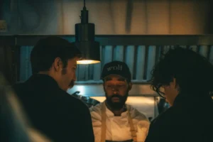 Three chefs gather closely in a dimly lit, moody kitchen environment illuminated by a single overhead pendant light. A centered chef wearing a baseball cap with the word "Wolf" looks down attentively alongside his colleagues.