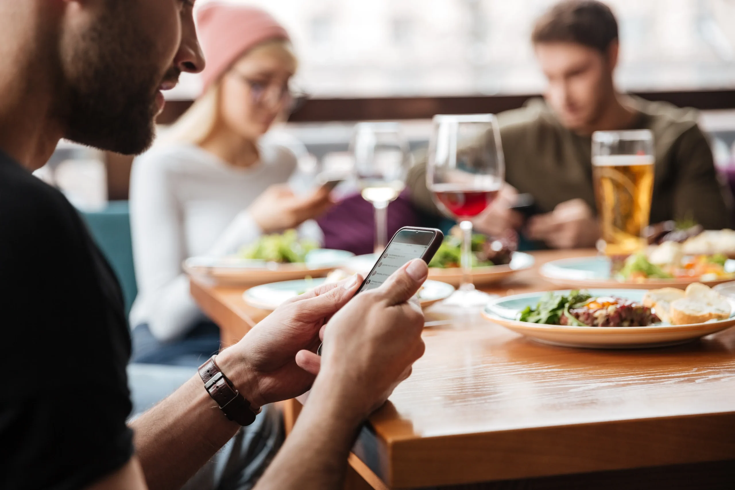 A group of friends sitting together at a restaurant table with food and drinks. Everyone is engrossed in their smartphones instead of interacting with each other.