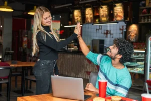 A smiling blonde woman stands next to a wooden table and gives a high five to a man seated in front of a laptop. They are inside a brightly lit restaurant with food, drinks, and cozy decor around them.