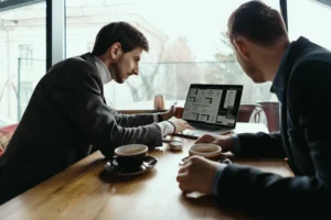 Two businessmen in suits sit across from each other at a wooden cafe table with coffee cups, reviewing a laptop screen. One man points toward the computer as they discuss work near a large window overlooking the city.