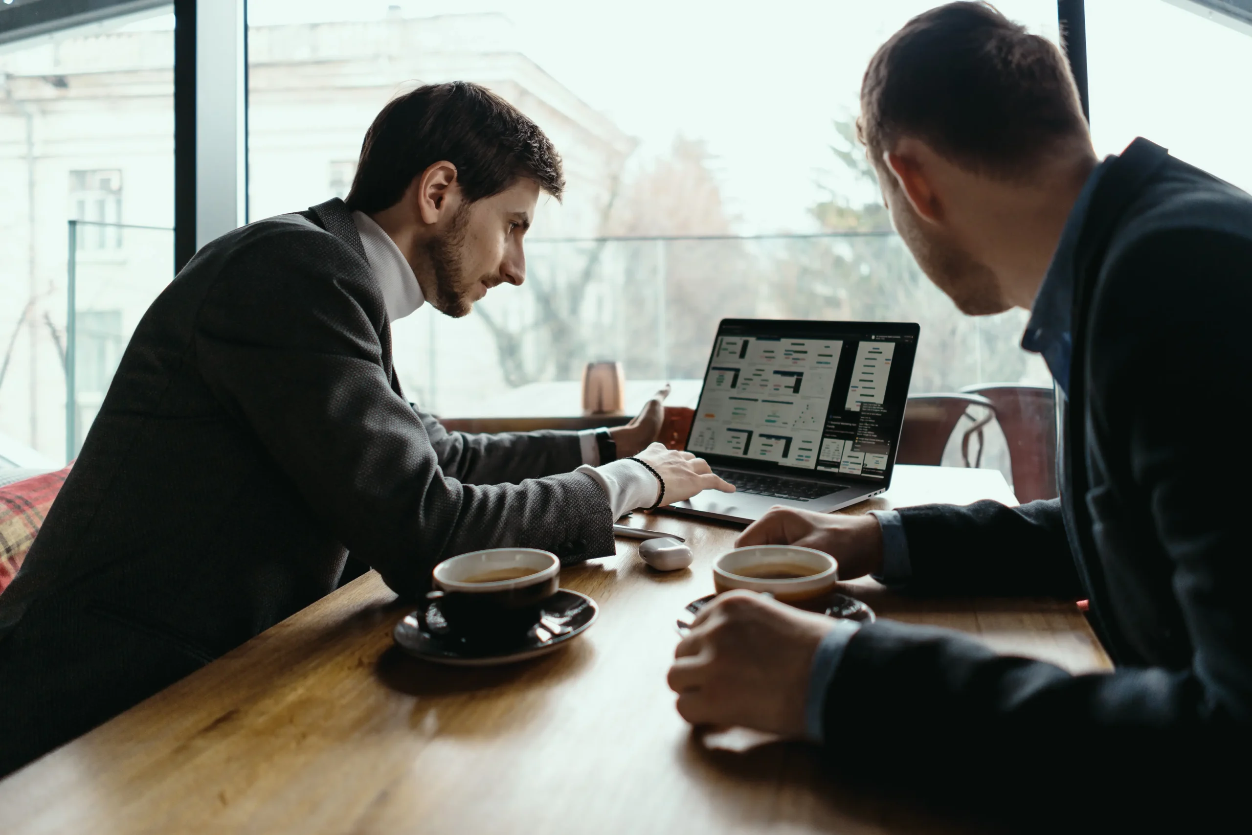 Two businessmen in suits sit across from each other at a wooden cafe table with coffee cups, reviewing a laptop screen. One man points toward the computer as they discuss work near a large window overlooking the city.