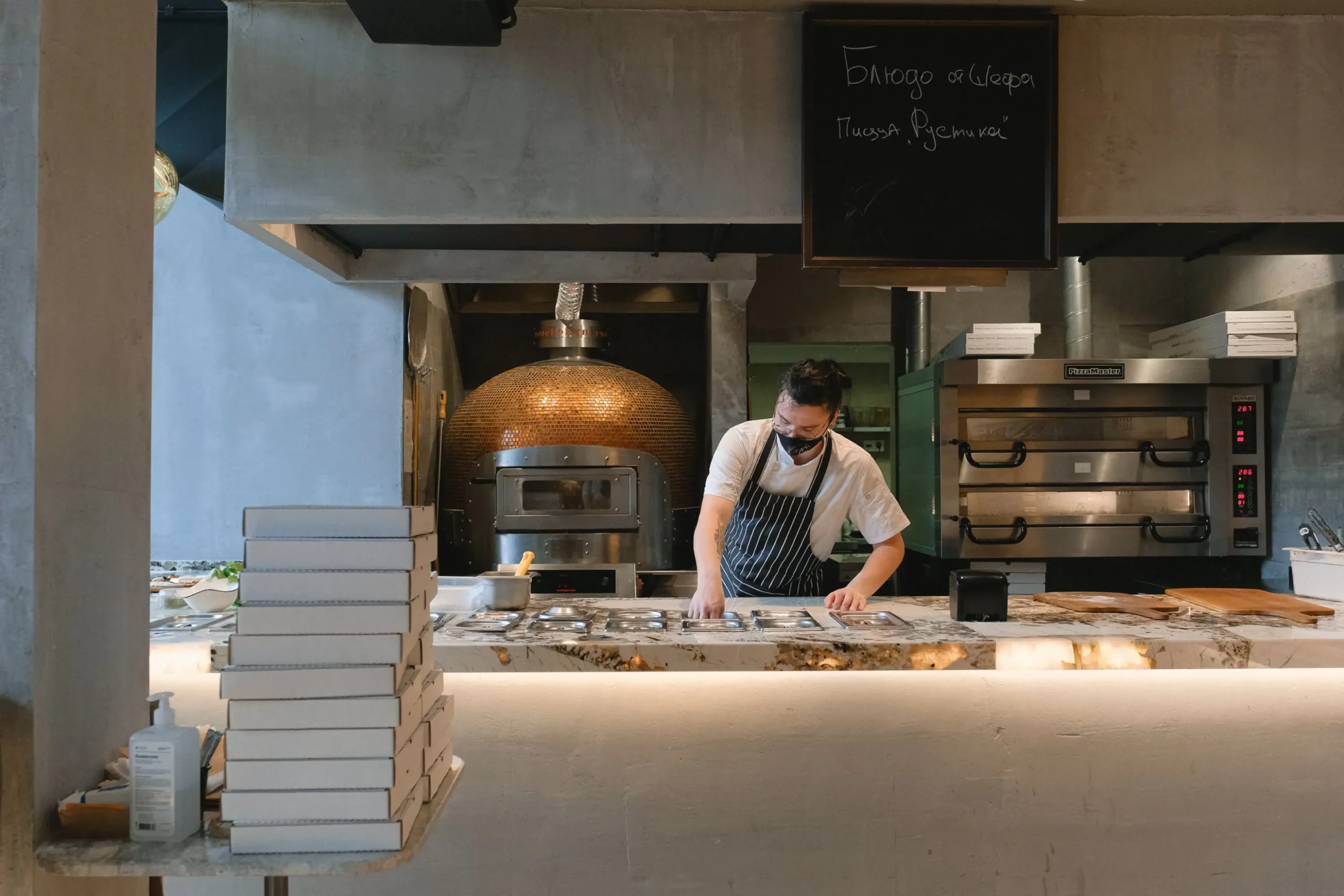A chef wearing a face mask and striped apron prepares food behind a marble counter in a modern pizzeria kitchen. A large dome pizza oven sits in the background, alongside stacked white pizza boxes in the foreground.