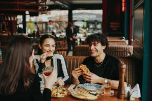Three young adults sit around a restaurant booth enjoying food, drinks, and casual conversation. A man with curly hair laughs while holding a sandwich, while a woman beside him smiles as she eats a tortilla chip.