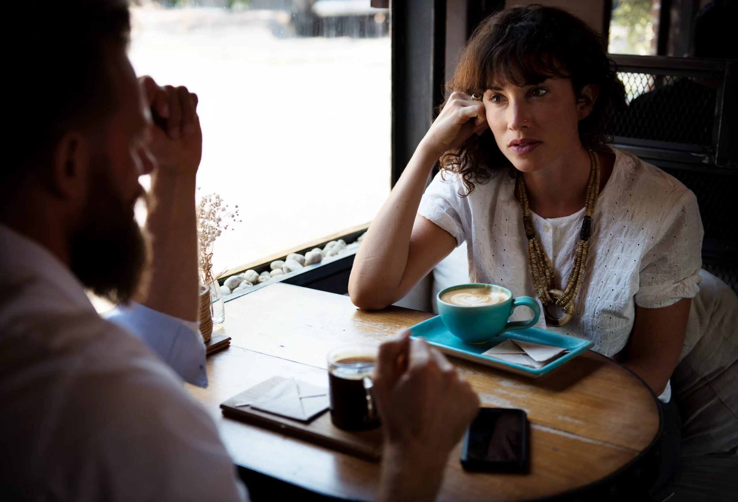 A woman with curly hair rests her head on her hand as she listens attentively to a man sitting across from her at a wooden coffee shop table. A teal mug, a glass of coffee, and small envelopes sit on the table between them in a cozy, sunlit space.