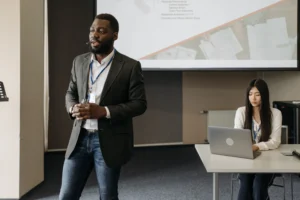 A presenter speaking into a microphone stands in front of a large screen displaying business process slides during a conference. An audience member on the right holds another microphone to ask a question as other attendees look on.