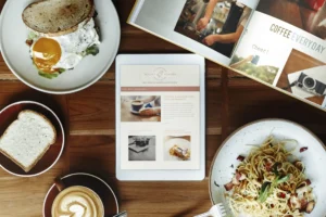 An overhead view shows a wooden table arranged with an open magazine, a coffee cup, a slice of bread, a plate of pasta, and an egg sandwich. A white tablet displaying a lifestyle blog sits directly in the center of the meal spread.