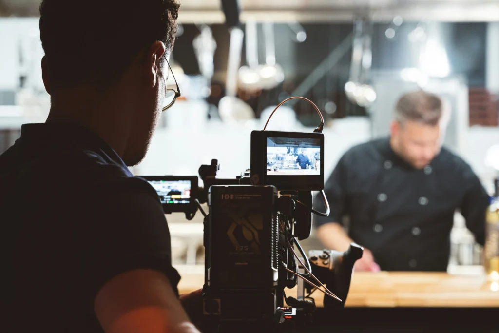 A videographer operates a professional camera while filming a chef in a modern commercial kitchen setting. The camera's monitor displays a clear, live feed of the chef working at a wooden counter.