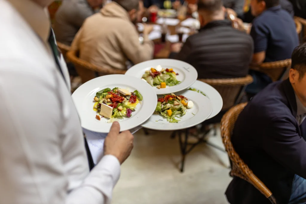 A waiter skillfully balances three white plates of fresh salad on one arm. In the background, seated guests gather around tables in a busy indoor dining area.