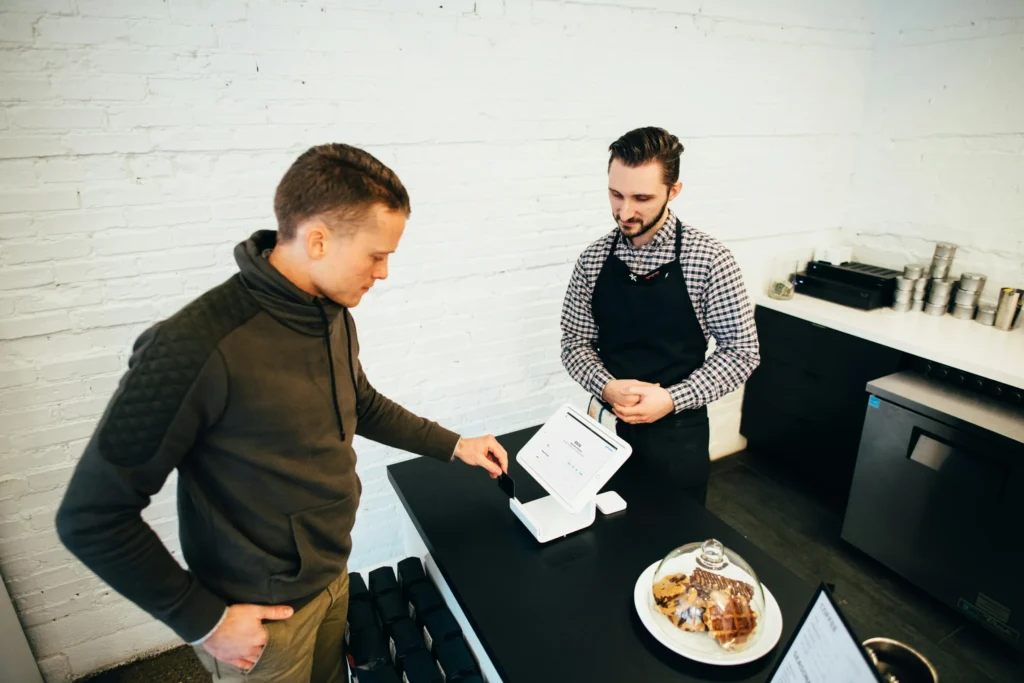 A customer in a grey pullover taps a credit card against a point-of-sale terminal at a coffee shop counter. Behind the counter, a barista wearing a black apron and plaid shirt watches the transaction.