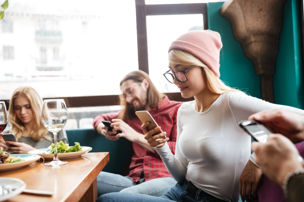 A group of young adults sits around a dining table at a restaurant, each focused intently on their own smartphone. Plates of food and glasses of wine rest on the table as the friends interact with their devices instead of each other.
