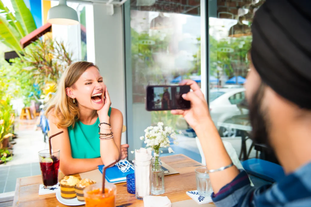 A man wearing a black turban takes a photo on his smartphone of a blonde woman wearing a green top who is laughing heartily at an outdoor café table. The table is set with drinks, muffins, and a small vase of flowers.