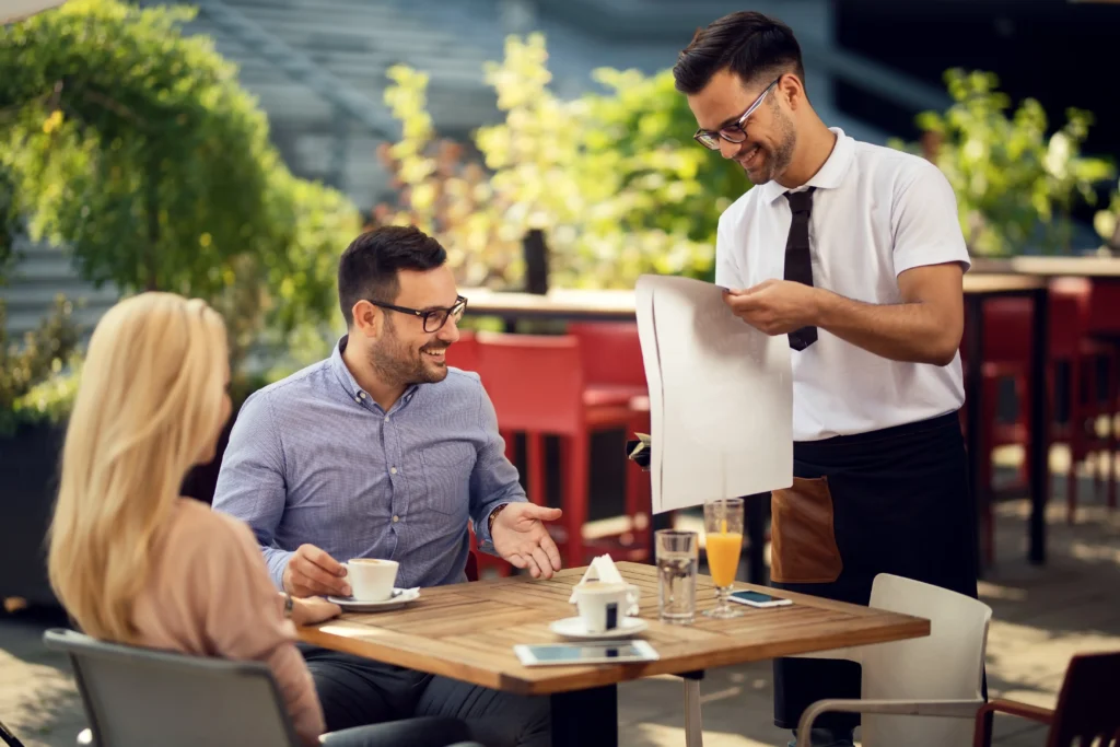 A smiling waiter in glasses laying down a paper placemat at an outdoor restaurant table. A seated couple enjoys coffee and chats cheerfully as he sets up their table.