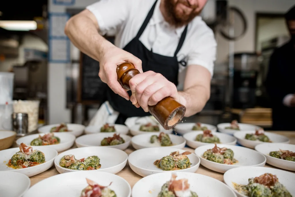 A chef wearing a black apron grinds fresh pepper over multiple small plates lined up on a counter. Each shallow bowl is filled with a green gnocchi or pasta dish topped with prosciutto and cheese.