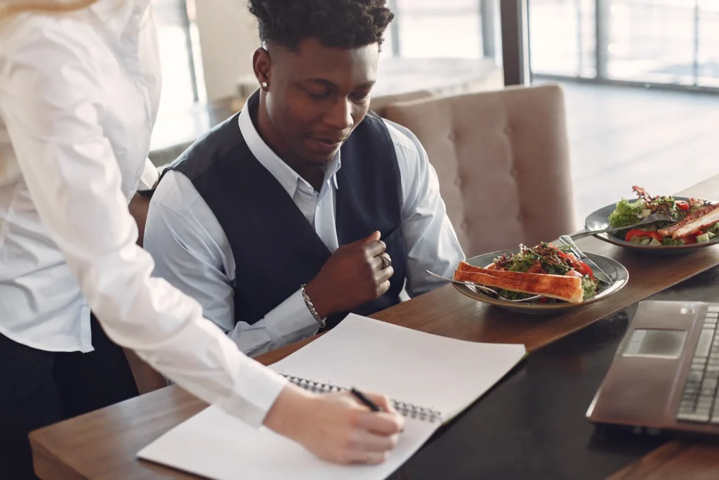A young man wearing a vest and collared shirt looks down attentively at a notepad on a wooden table. A colleague stands beside him, writing in the notebook during a business meeting over lunch.