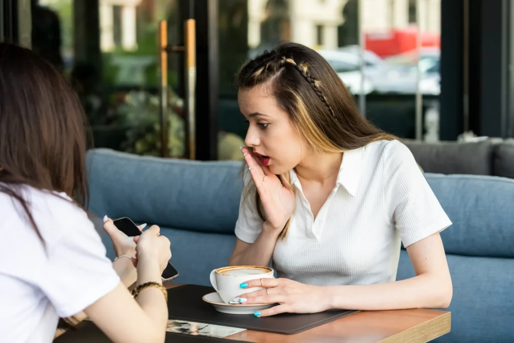 A young woman sits at a cafe table with a cup of coffee, holding her hand up to her mouth with a shocked, open-mouthed expression. She looks intently at a smartphone held by her friend sitting across from her.