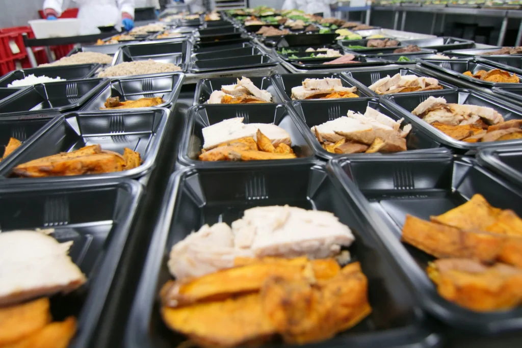 Rows of black plastic containers are arranged on kitchen tables, filled with sliced chicken and roasted sweet potatoes. In the background, workers in white coats and gloves assemble prepped meal-prep boxes on a catering line.
