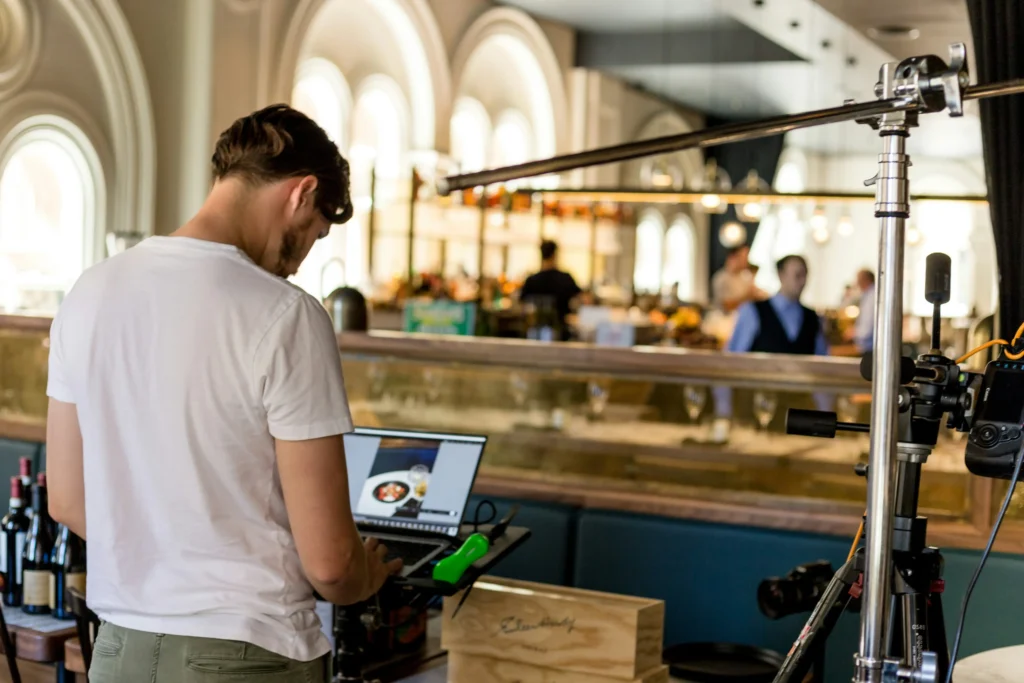 A man wearing a white t-shirt stands behind a laptop reviewing photos inside an upscale restaurant or bar. Professional camera equipment on a tripod is set up next to him during the photo shoot.