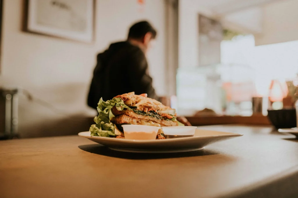 A stacked sandwich with fresh lettuce and two small dipping sauce cups sits on a plate atop a wooden table. In the softly blurred background, a person stands inside a cozy café setting.