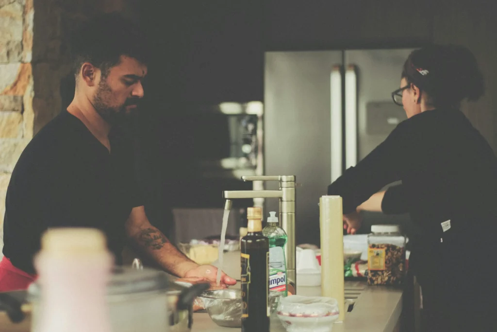 A man and a woman prepare a meal together at a modern kitchen counter. The man washes his hands under a running faucet while the woman works alongside him near cooking ingredients and supplies.