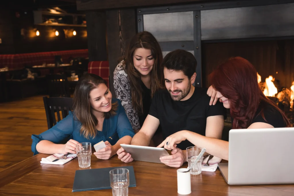 Four young friends gather around a wooden table in a dimly lit restaurant, smiling as they look closely at a digital tablet together. A laptop, drink glasses, and smartphones sit nearby on the table next to a cozy, lit fireplace in the background.
