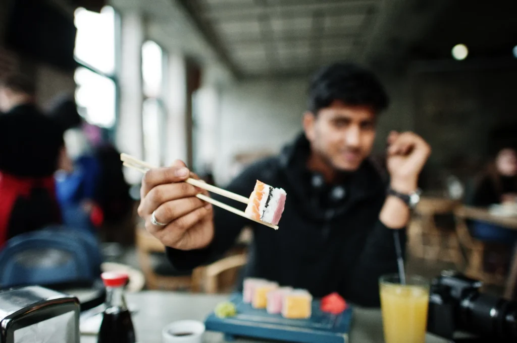 A close-up shot focuses on a hand holding a piece of sushi with wooden chopsticks in front of the camera. In the softly blurred background, a young man sits at a cafe table with a tray of sushi and a drink.
