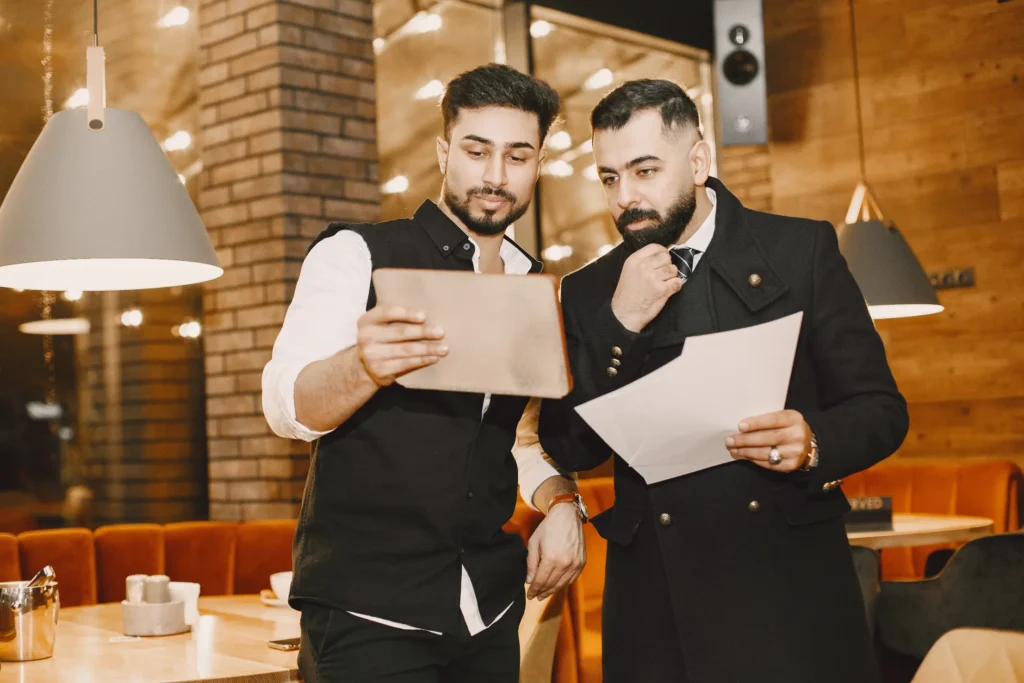 Two well-dressed men stand together in a warmly lit restaurant while focused on reviewing documents. One holds up a tablet or menu folder, while the other holds sheets of paper with a thoughtful expression.