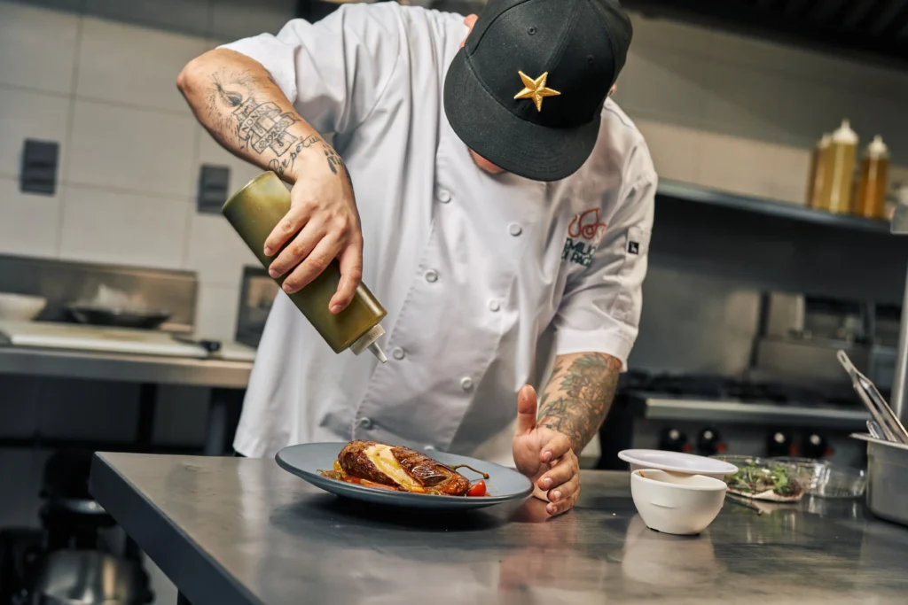 A chef wearing a white jacket and a black baseball cap carefully drizzles sauce from a green squeeze bottle onto a plated dish. The tattooed chef works on a metal kitchen counter inside a modern restaurant kitchen setup.