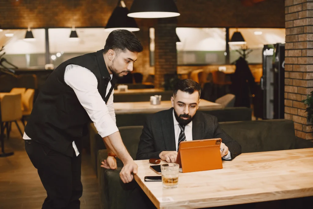 A man in a suit sits at a wooden table in a restaurant, working on a tablet with a brown case. Another man wearing a vest and white shirt leans over the table to look at the screen with him.