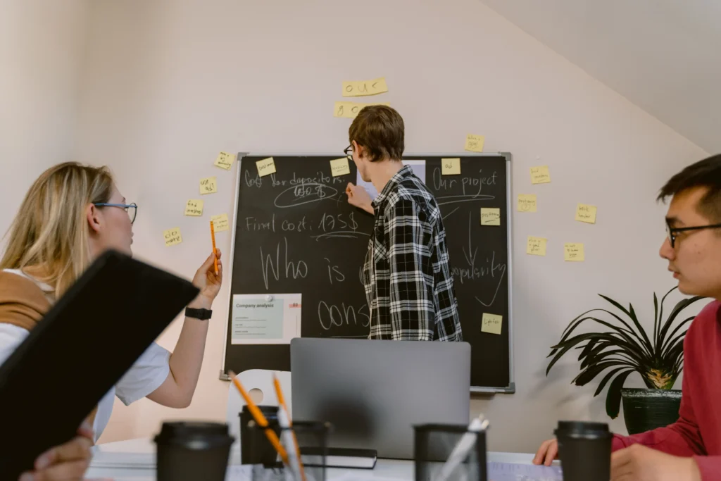 A man in a plaid shirt writes notes on a blackboard during a collaborative team strategy session. Colleagues sit at a table around him with laptops, notebooks, and coffee cups while sticky notes dot the board and wall.