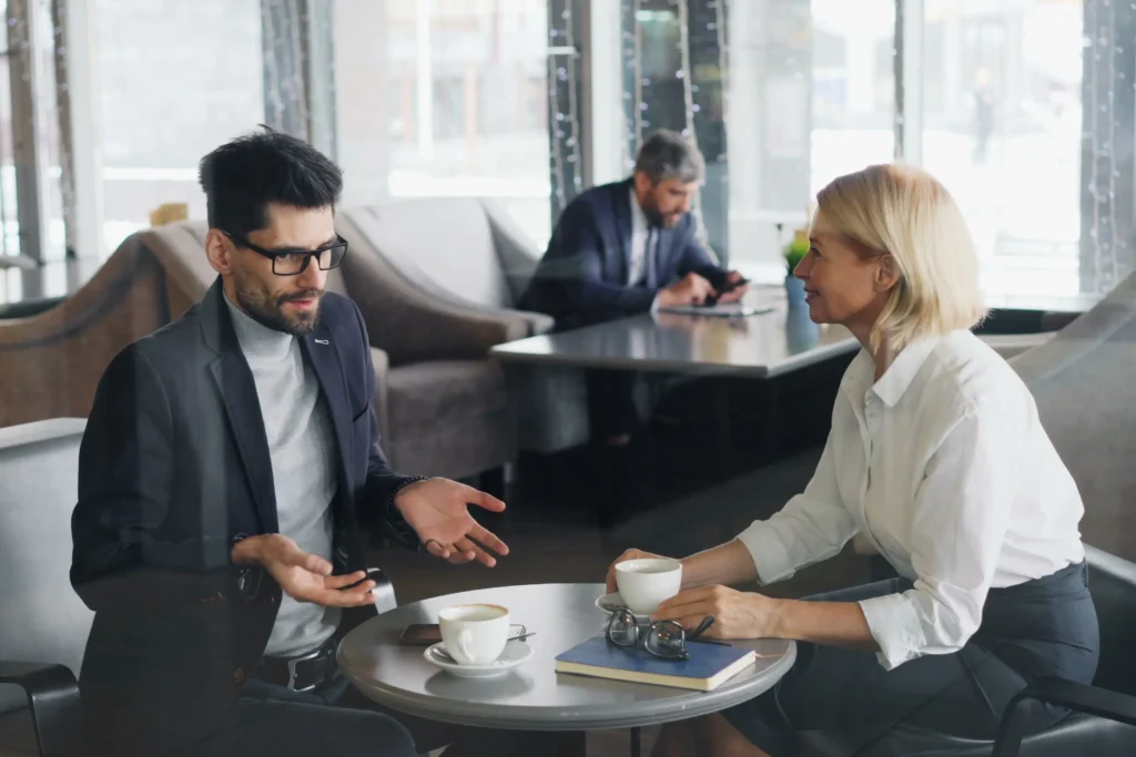 A man in a suit jacket and glasses gestures with open hands while conversing with a blonde woman across a small table in a bright modern cafe. Coffee cups, a notebook, and eyeglasses sit between them as they engage in their business discussion.