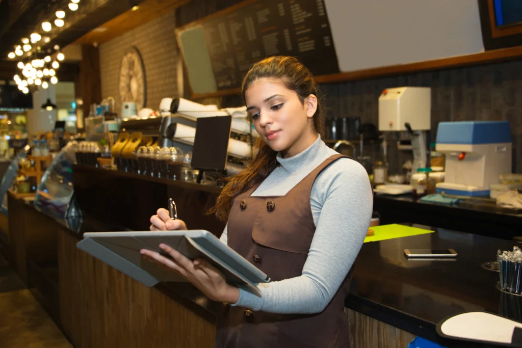 A young female cafe worker wearing an apron writes on a tablet while standing behind the counter. The modern cafe background features a warm, warm-lit aesthetic with coffee equipment visible behind her.