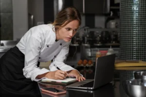 A female chef wearing a white chef coat leans over a kitchen counter while focused on her laptop screen. She holds a pen to take notes in a notebook open next to her device, with ingredients and kitchen gear in the background