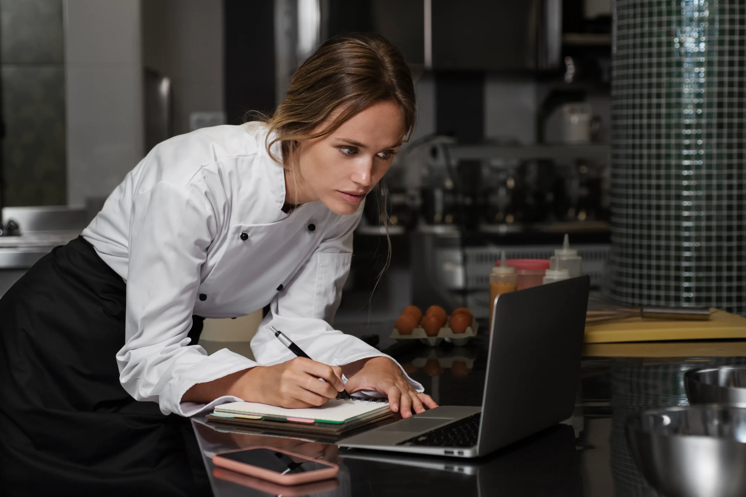 A female chef wearing a white chef coat leans over a kitchen counter while focused on her laptop screen. She holds a pen to take notes in a notebook open next to her device, with ingredients and kitchen gear in the background